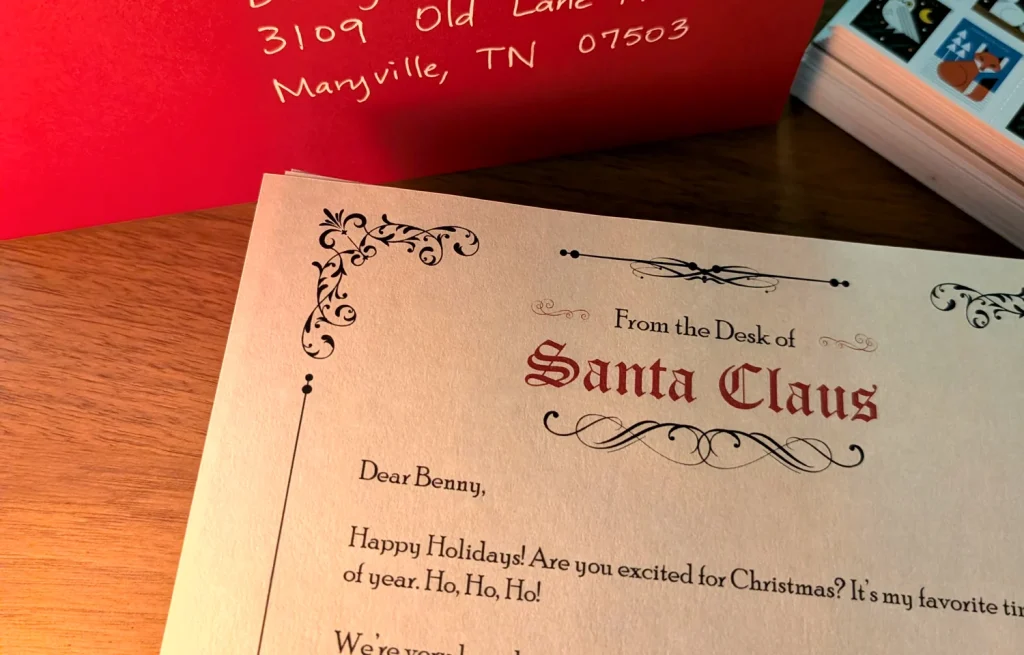 A printed letter from Santa Claus reading Dear Benny, Happy Holidays, beside the red envelope hand-addressed to the child and a sheet of stamps