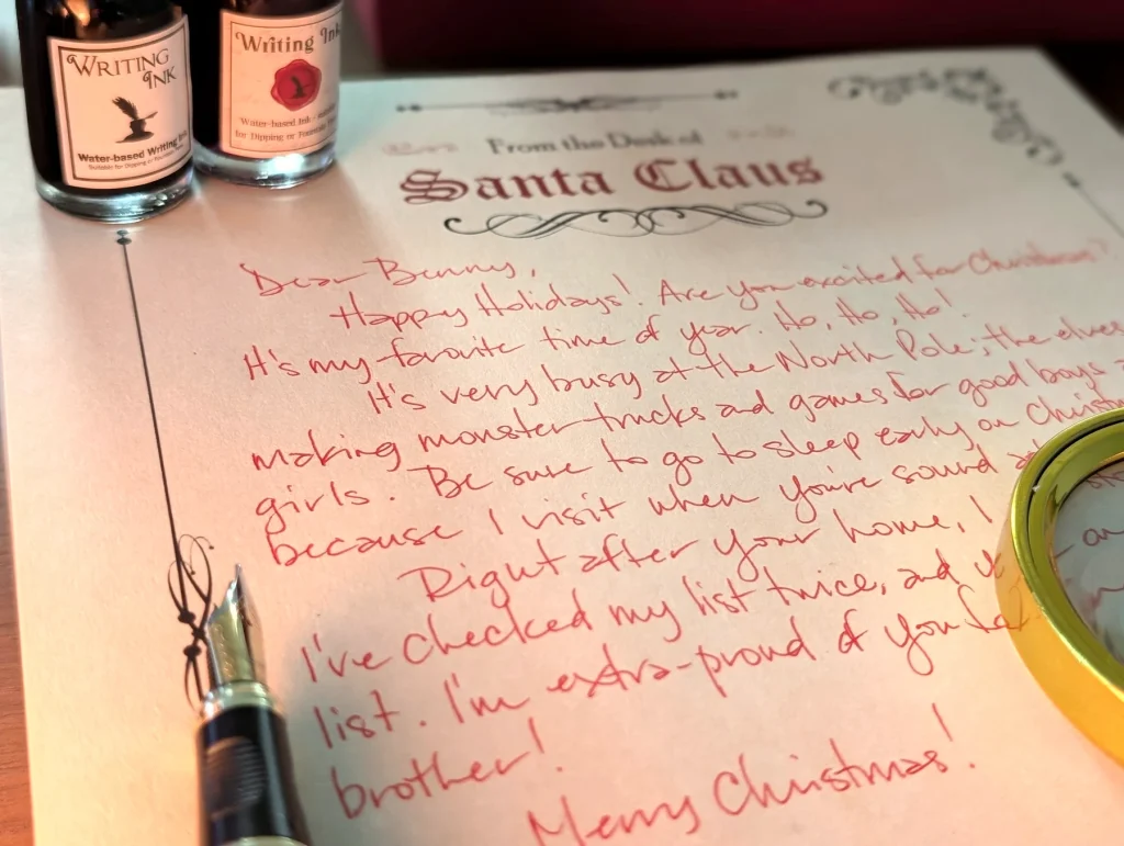 A full letter from Santa handwritten in red ink on Santa Claus letterhead, photographed close up with two bottles of writing ink, a fountain pen and a magnifying glass