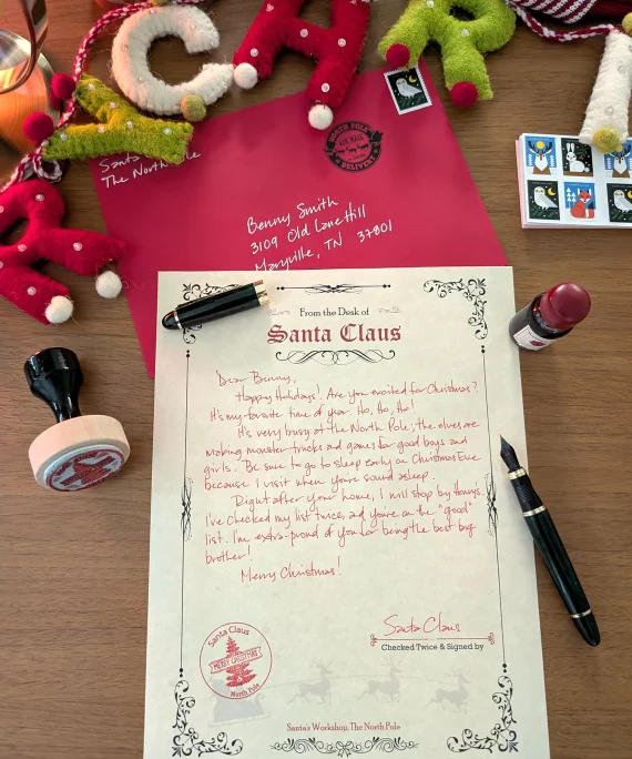 Handwritten Santa letter flat lay with the red envelope, magnifying glass, real stamps and the Merry Christmas felt garland