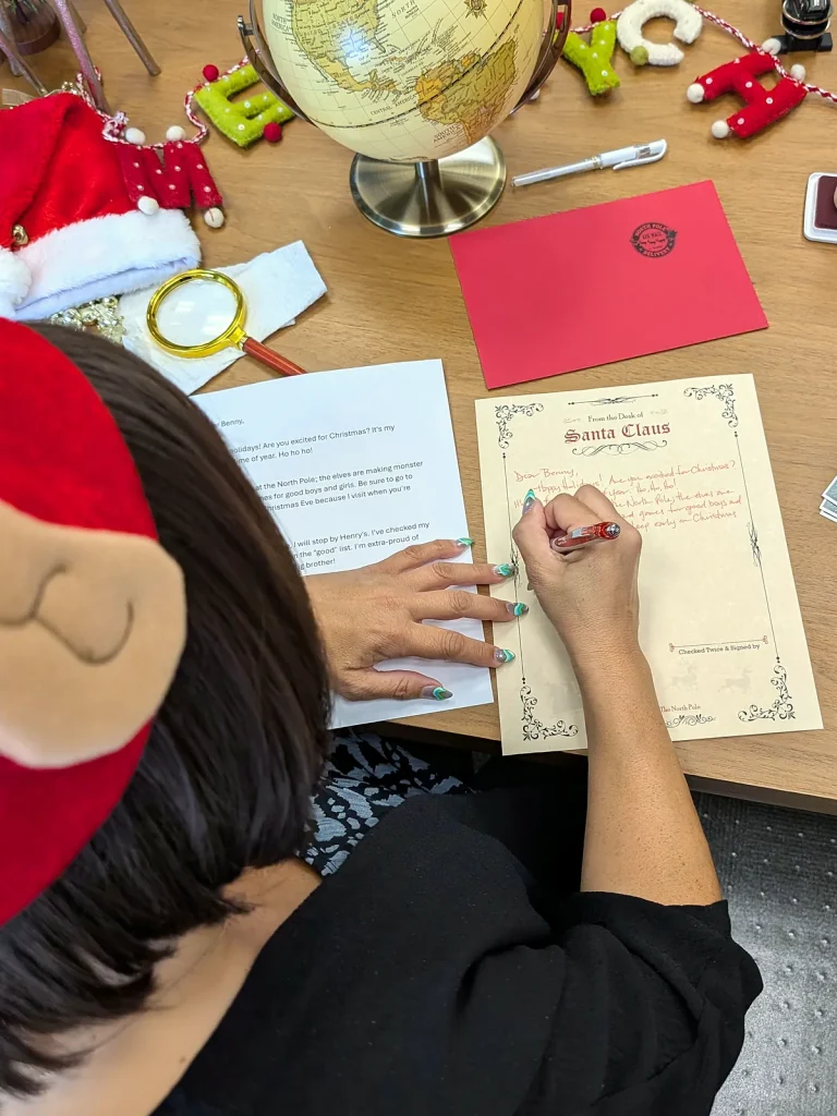 Writing a Santa letter at the desk in an elf hat, seen from above, with the globe, the red envelope and the stamps