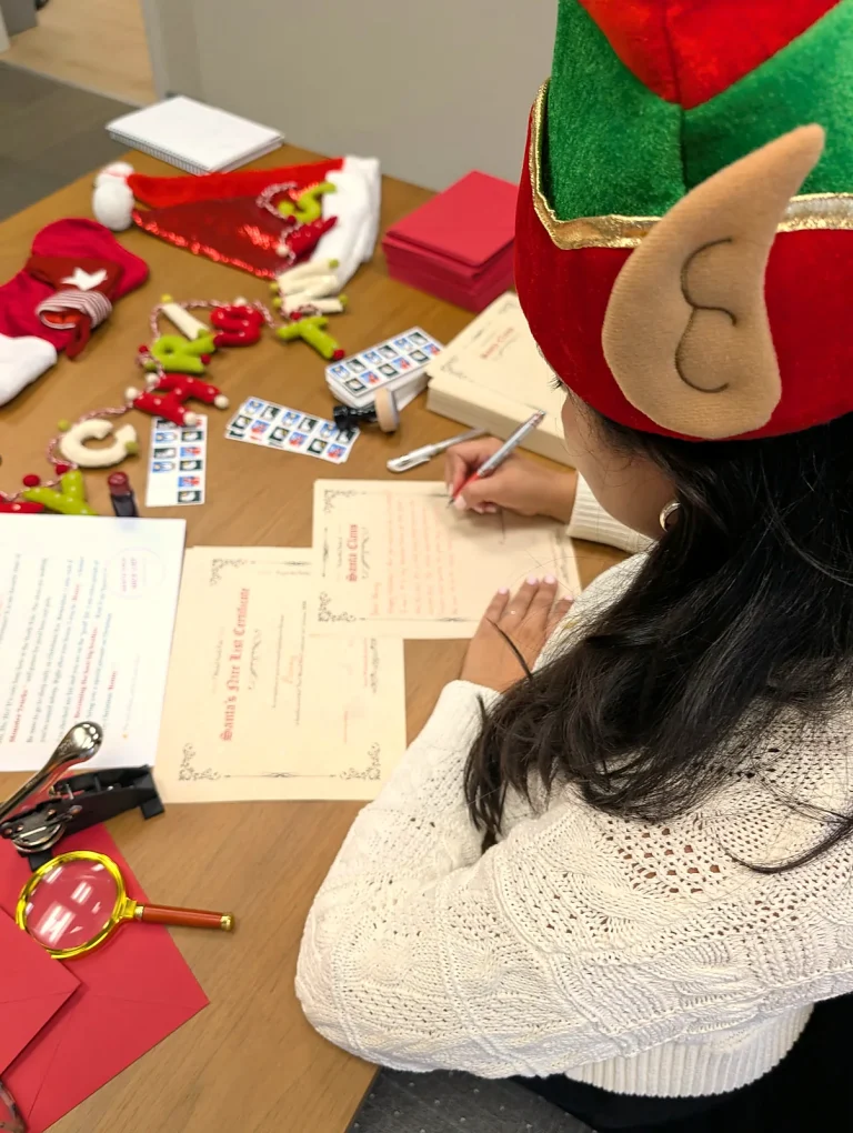 Writing Santa letters at the desk in an elf hat, seen from behind, with the stockings and the Merry Christmas garland